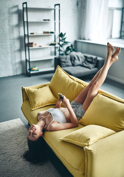 Good morning! Attractive young woman is having rest at home. Girl is listening music while lying on sofa. - Foto, afbeelding