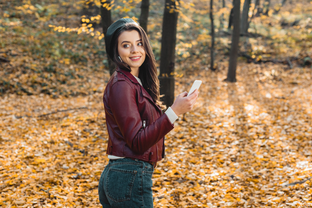 cheerful fashionable girl in beret and leather jacket holding smartphone and looking away outdoors  - Foto, Imagem