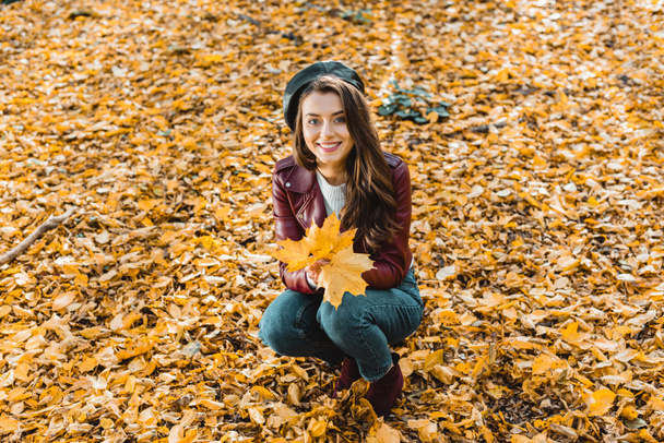 high angle view of happy fashionable girl in beret and leather jacket sitting with yellow in autumnal forest - Фото, изображение