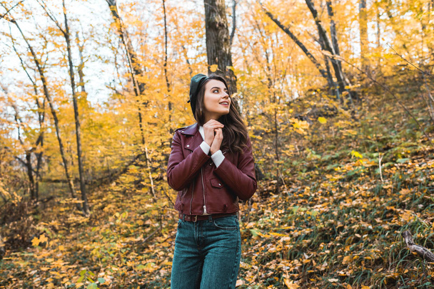 low angle view of fashionable young woman in beret and leather jacket looking away in forest  - Photo, Image