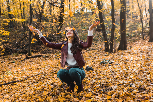 lächelnde attraktive Frau in stylischer Lederjacke, die Spaß mit gelben Blättern im herbstlichen Wald hat  - Foto, Bild
