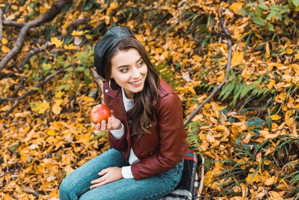 vista ad alto angolo del modello femminile in giacca di pelle seduta su coperta e tenuta mela rossa nella foresta autunnale
 - Foto, immagini
