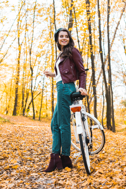low angle view of happy female model in beret and leather jacket posing near bicycle in autumnal forest - Foto, Bild