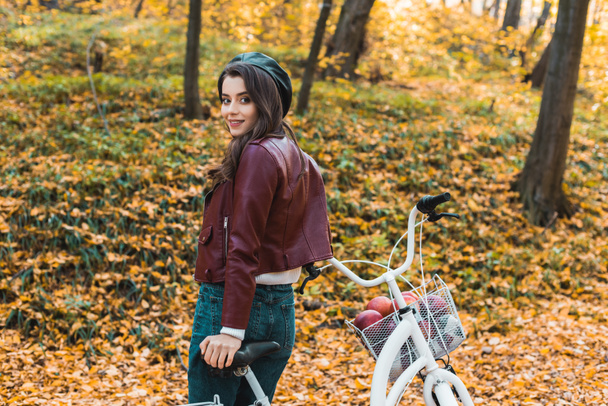 selektiver Fokus des modischen weiblichen Modells in Baskenmütze und Lederjacke, das in der Nähe von Fahrrädern im herbstlichen Wald posiert - Foto, Bild