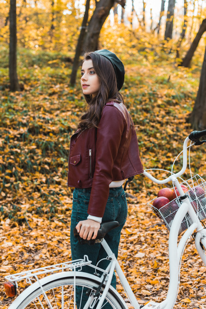attractive stylish girl in beret and leather jacket posing near bicycle in autumnal forest - Photo, Image