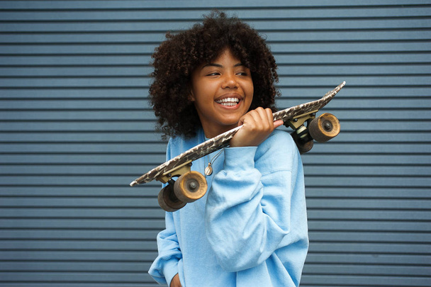 Closeup portrait of smiling girl with natural beauty wearing blue sweatshirt holding her cool skateboard on a shoulder on an urban setting background at daytime on a lovely day. - Foto, Imagen