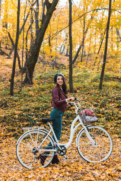 mulher na moda feliz na jaqueta de couro elegante e boina transportando bicicleta na floresta
 - Foto, Imagem