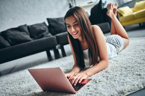 Good morning! Attractive young woman is having rest at home. Girl lying on a carpet with laptop. - Foto, imagen