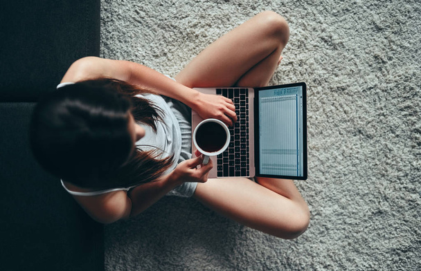 Dzień dobry! Atrakcyjna młoda kobieta jest o odpoczynku w domu. Widok z góry z dziewczyna siedzi na dywanie filiżankę kawy i laptopa. - Zdjęcie, obraz