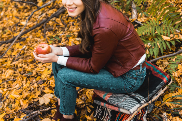 vue partielle de la fille élégante en veste en cuir assis sur la couverture et tenant pomme rouge dans la forêt automnale
 - Photo, image