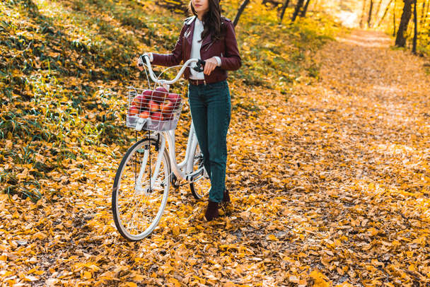 partial view of girl in stylish leather jacket and beret carrying bicycle in yellow autumnal forest - Fotó, kép