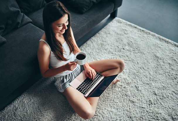 Dzień dobry! Atrakcyjna młoda kobieta jest o odpoczynku w domu. Siedząc na dywanie filiżankę kawy i laptopa. - Zdjęcie, obraz
