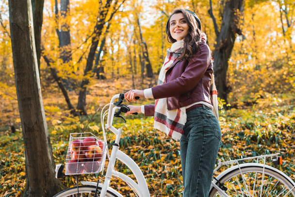 low angle view of fashionable girl in leather jacket and beret carrying bicycle in forest - Photo, Image