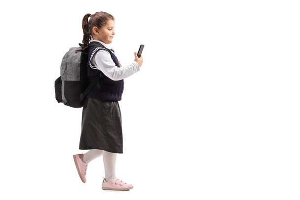 Full length shot of a little schoolgirl in a uniform using a mobile phone and walking isolated on white background - Fotoğraf, Görsel