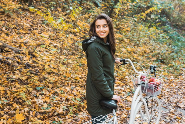 smiling young woman carrying bicycle with basket full of apples in yellow autumnal forest  - Foto, Imagem
