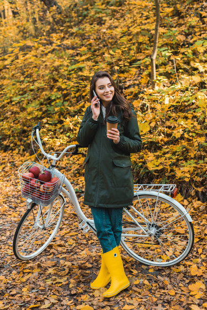 smiling young woman with paper coffee cup talking on smartphone near bicycle in autumnal forest  - Photo, image