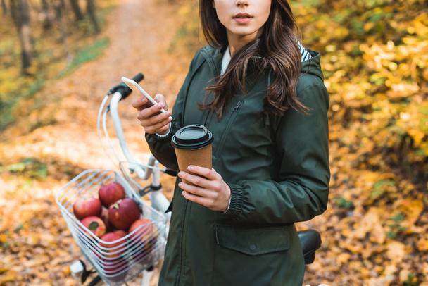 przycięty obraz kobiety z kubkiem kawy jednorazowe za pomocą smartfona w pobliżu rower na zewnątrz  - Zdjęcie, obraz