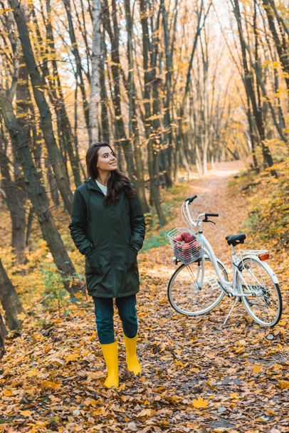 chica feliz en botas de goma amarillas caminando cerca de la bicicleta en el bosque otoñal
 - Foto, Imagen