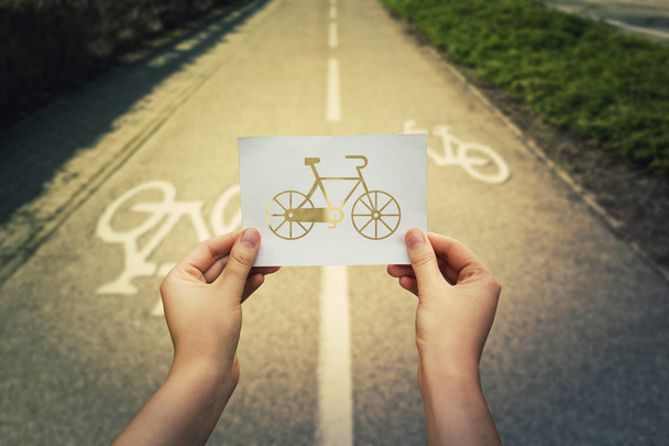 Close up of woman hands holding a white paper sheet with bicycle icon over bike lane background. Healthy life rhythm doing sport. - Foto, afbeelding