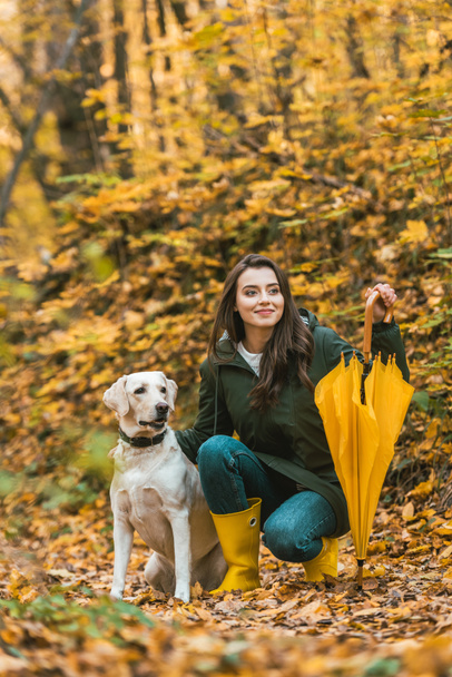 happy young woman with yellow umbrella siting with dog in autumnal forest  - Fotografie, Obrázek