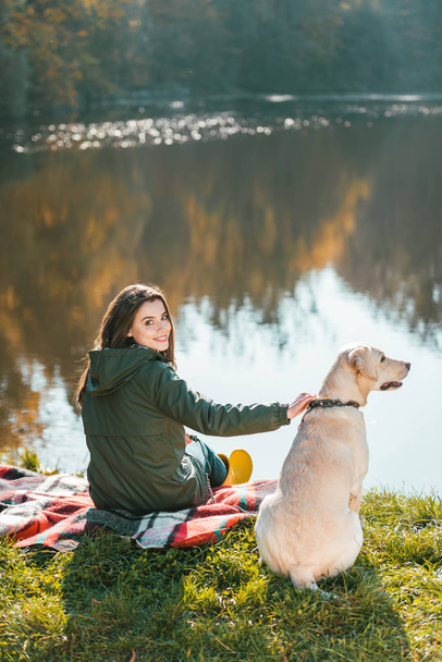 smiling young woman siting on blanket with adorable golden retriever near pond in park - Photo, Image