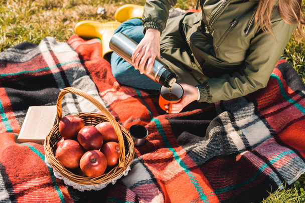 partial view of woman pouring coffee from thermos into cup while sitting on blanket with basket full of apples outdoors  - Fotografie, Obrázek