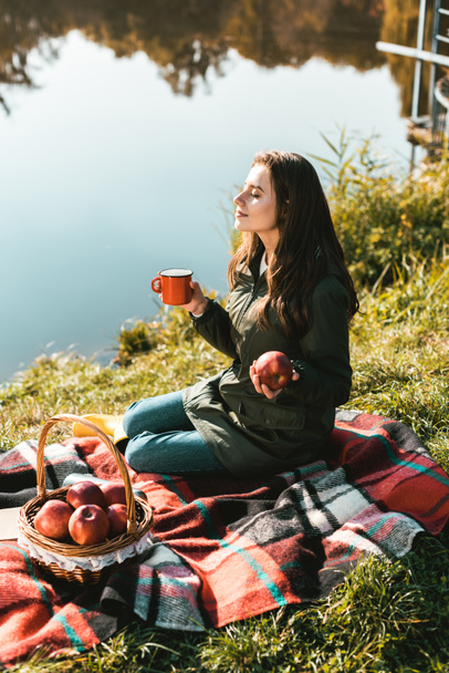 χαρούμενο κορίτσι με μήλο και φλιτζάνι καφέ, κάθονται σε κουβέρτα κοντά σε λίμνη  - Φωτογραφία, εικόνα