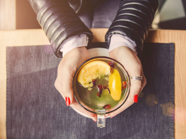 Female hands with red manicure holding cup of tea with herbs and lemon on table in cafe. - Photo, Image