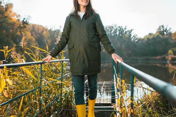 vista parcial de mujer joven con chaqueta y botas de goma amarillas posando cerca del estanque en el parque
 - Foto, imagen