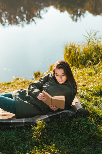 high angle view of young woman reading book while laying on blanket near pond in park - Fotografie, Obrázek