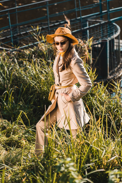 stylish female model in sunglasses, trench coat and hat posing near grass - Photo, Image