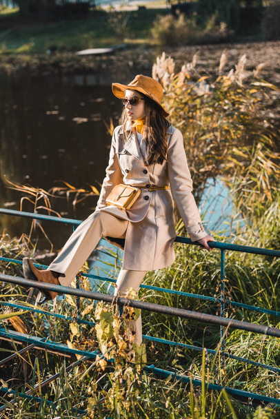fashionable woman in sunglasses, trench coat and hat sitting on railing near pond  - Fotografie, Obrázek
