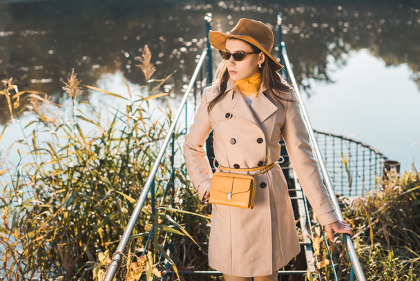 foco seletivo da mulher na moda em óculos de sol, casaco de trincheira e chapéu posando perto da lagoa no parque
 - Foto, Imagem