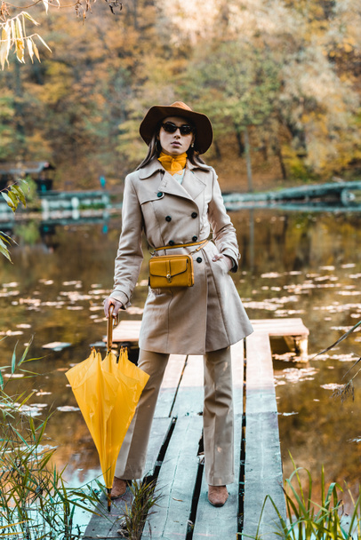 confident stylish woman in sunglasses, trench coat and hat posing with yellow umbrella near pond in park - Photo, Image