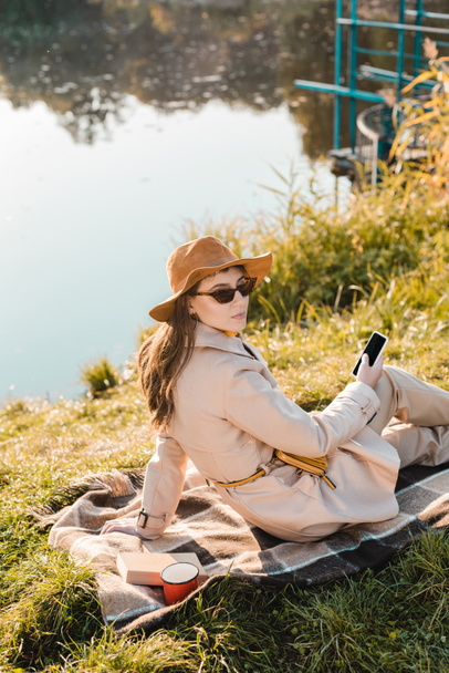 όμορφη κομψή γυναίκα με καπέλο και γυαλιά ηλίου κρατώντας smartphone και να κάθεται στην κουβέρτα κοντά Λίμνη στο πάρκο - Φωτογραφία, εικόνα