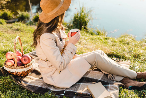 Seitenansicht einer jungen Frau in Trenchcoat und Hut, die eine Tasse Kaffee hält, während sie auf einer Decke im Freien sitzt  - Foto, Bild