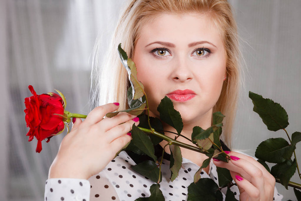 Romance, valentine day gifts concept. Beautiful blonde young woman holding red rose near face looking melancholic - 写真・画像