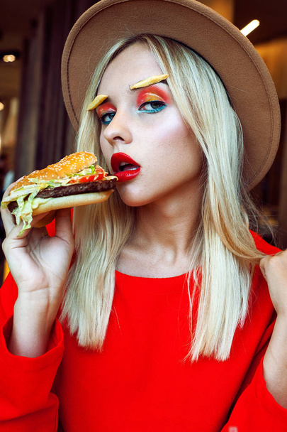 Young beautiful blonde teenage girl in red with bright creative makeup wearing hat holding hamburger at her mouth while looking at camera at restaurant. Unhealthy diet concept. - Foto, imagen