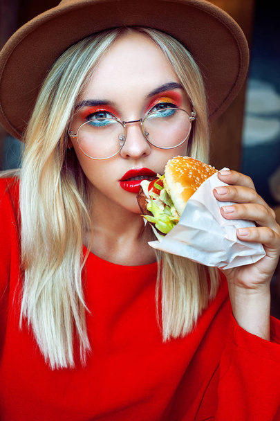 Young beautiful blonde teenage girl in red with bright creative makeup wearing hat and glasses holding hamburger close to her face. Looking at camera. Unhealthy diet concept. - 写真・画像