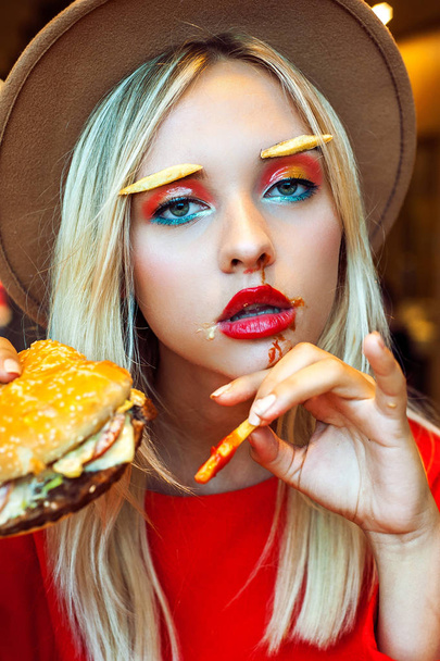 Beautiful teenage girl in red and a hat wearing bright creative makeup and fries instead brows eating fast food with a ketchup on her face at the restaurant. Looking at camera. Unhealthy diet concept. - 写真・画像