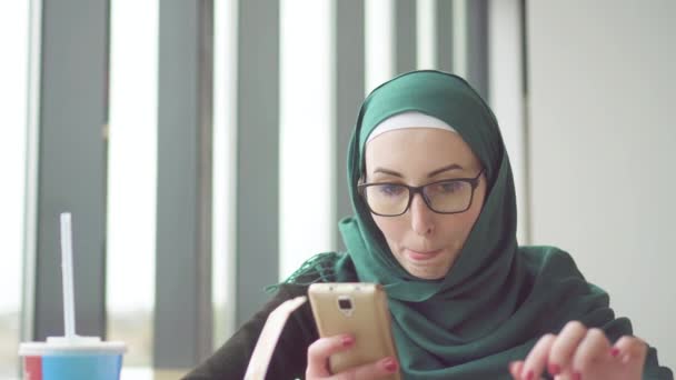 Young muslim woman in a hijab is sitting at a table by the window using the phone and eating potatoes - Felvétel, videó