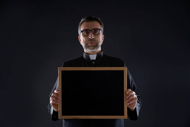 Priest holding empty blackboard copy space isolated on black - Zdjęcie, obraz
