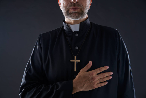 Priest hand in heart gesture with cross on black background - Photo, image