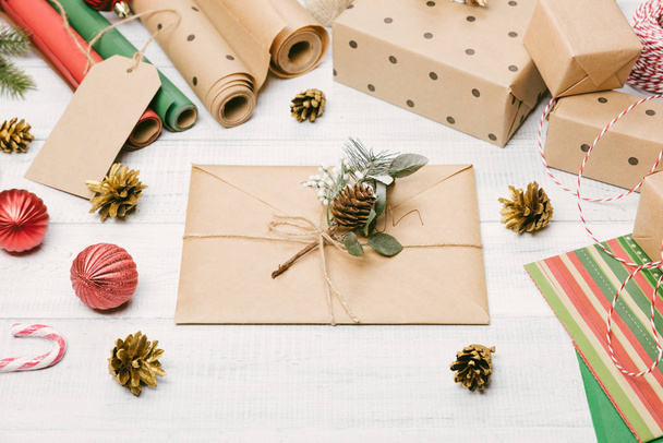 クラフト紙に包まれたクリスマスプレゼントやギフトボックス、木製のテーブルに封筒、モミの木の枝や装飾、トップビュー。フラットレイアウト、テキストのコピースペース. - 写真・画像