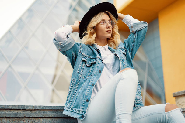 Close-up portrait of interesting woman in glasses touching her hat on urban background. Photo of fashionable girl with beautiful blonde hair posing while sitting on stairs. - 写真・画像