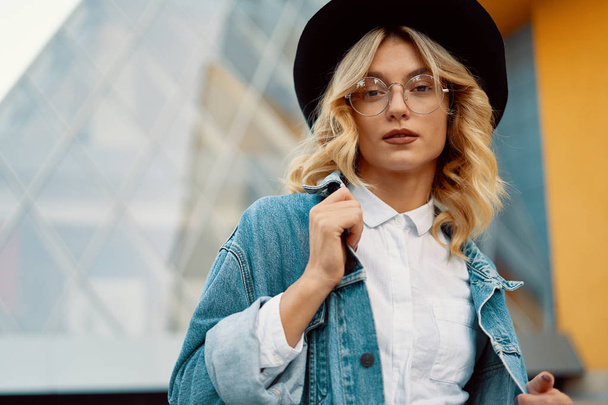 Close-up portrait of a cheerful white woman in glasses touching her jacket collar on urban background. Photo of fashionable girl with beautiful blonde hair posing for the camera. - Photo, Image