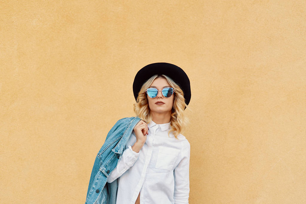 Porträt eines Hipster-Mode-Mädchens, das auf einer Schulter vor dem gelben Wandhintergrund steht. Youth Urban Fashion Konzept. Kopierraum. - Foto, Bild