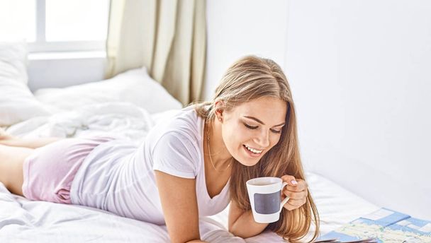 glückliches junges Mädchen im Bett mit Handy, Tablet und Karten auf der Suche nach Reisen - Foto, Bild
