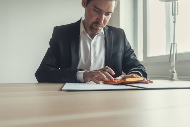 Imagem retrô de um jovem empresário com barba usando calculadora enquanto se senta em sua mesa de escritório revisando documentos
. - Foto, Imagem