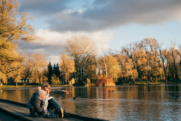 Mother and son sitting near lake in autumn park on sunset and looking away. Family having fun, hugging, laughing, relaxing, enjoy life.Close up sunny lifestyle fashion portrait of happy stylish family - Foto, afbeelding